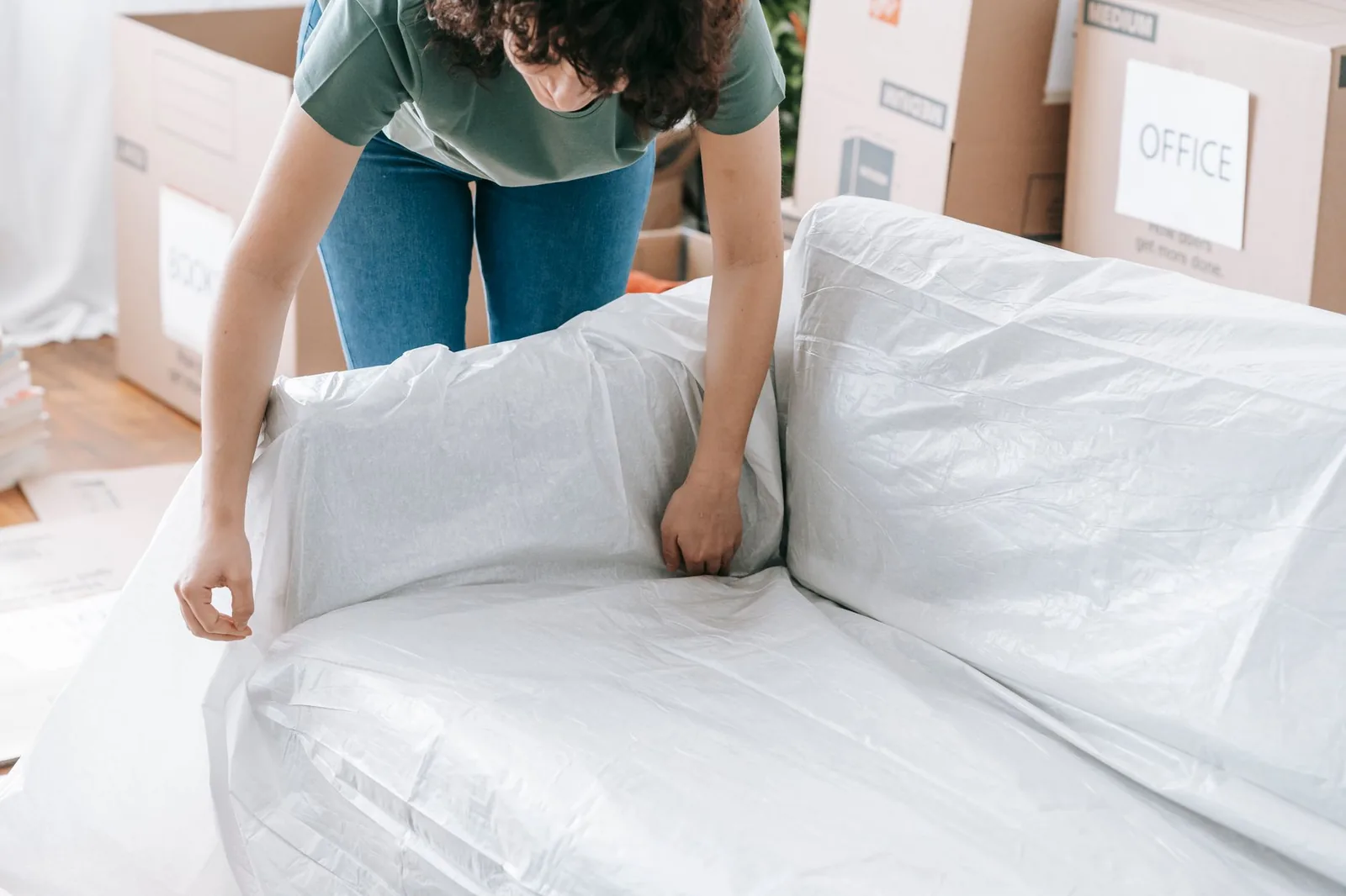 Una donna avvolge un divano in un telo di plastica bianco, con gli scatoloni del trasloco alle spalle