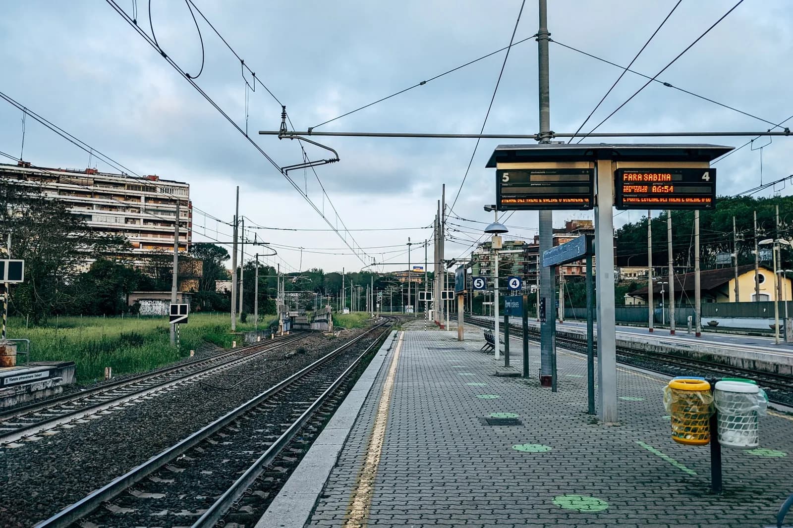 La banchina vuota di una stazione ferroviaria all'alba, con i binari e i palazzi sullo sfondo.