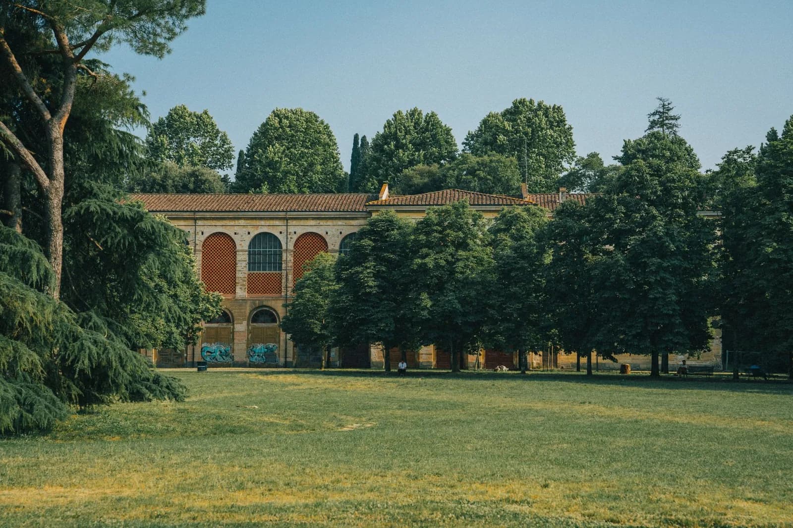 Un ampio prato con alberi alti e, sullo sfondo, un edificio ad arcate rosse in un parco di Firenze.